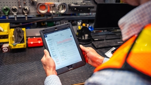Worker in a high-vis vest holding a tablet showing a data dashboard in a workshop with tools visible behind