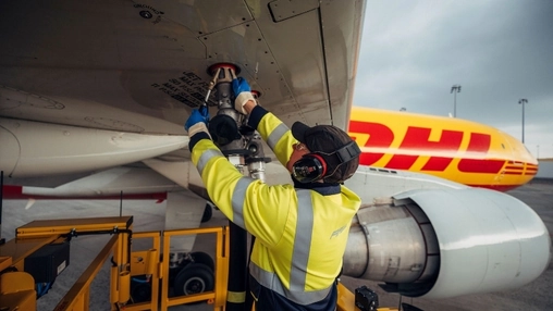 Airport worker in yellow high-vis and ear defenders fuelling a white aircraft with a DHL cargo plane behind