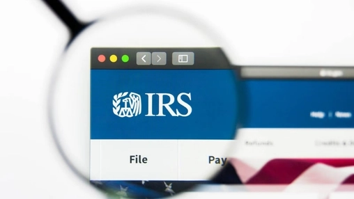 A hand holds a magnifying glass over a monitor displaying the IRS website with file and pay tabs shown